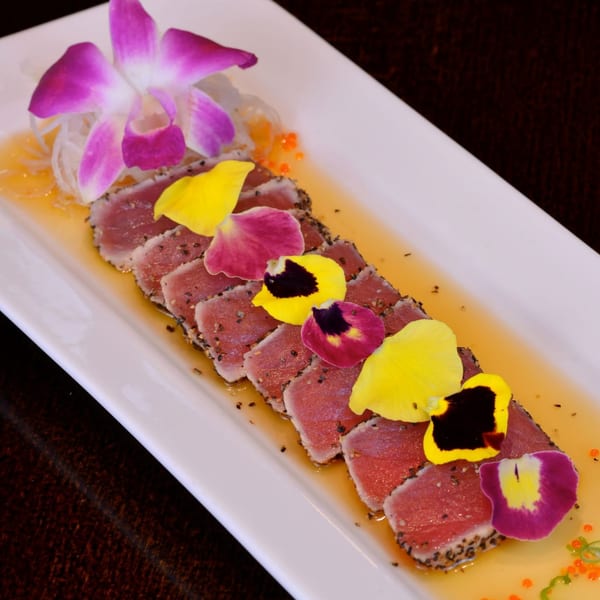 Seared tuna slices with sesame seeds, edible flowers, and a purple orchid on a white rectangular plate.